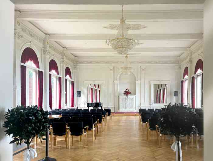 Der hell gestrichene Spiegelsaal ist Trauort im Alten Kurhaus Bad Zwischenahn. An der Decke üppige Kristallleuchter und rote Vorhänge an den Fenstern. Der Saal ist großzügig bestuhlt.